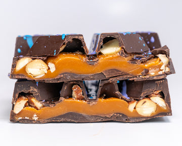 A close-up of a broken chocolate bar stacked on top of another, showing dark chocolate, caramel , sea salt and almonds with colorful cocoa butter splatters